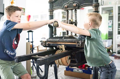 Zwei Jungs testen den Bilddruck mit einer händischen Maschine im KunstMuseum Donau-Ries.