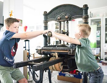 Zwei Jungs testen den Bilddruck mit einer händischen Maschine im KunstMuseum Donau-Ries.