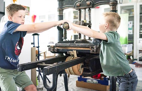 Zwei Jungs testen den Bilddruck mit einer händischen Maschine im KunstMuseum Donau-Ries.