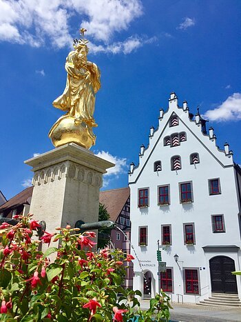 Marktplatz Wemding Hoch aufragende Fassade des historischen Rathaus mit einem blauen Himmel im Hintergrund. Im Vordergrund steht eine goldene Marienstatue mit Brunnen.