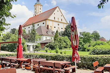 Im Vordergrund sieht man einen Teil des Biergartens mit seinen Tischen und Bänken. Im Hintergrund sieht man Wallfahrtsbasilika Maria Brünnlein.