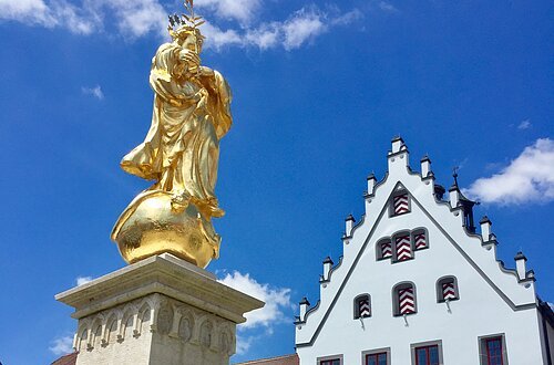 Marktplatz Wemding Hoch aufragende Fassade des historischen Rathaus mit einem blauen Himmel im Hintergrund. Im Vordergrund steht eine goldene Marienstatue mit Brunnen.