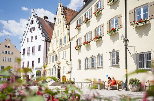 Zwei Freundinnen sitzen auf einer Bank vor dem Verwaltungsgebäude am historischen Marktplatz und unterhalten sich.